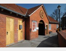 Backsteingebäude mit rotem Ziegeldach, einem rundbogigen Eingangsbereich mit der Aufschrift „Spielplatz“ und einem dekorativen goldfarbenen Zifferblatt über dem Eingang. Links befinden sich zwei Holztüren mit diagonalem Muster, rechts Laternenpfähle und Bäume im Hintergrund.