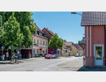 Straßenszene in einem Wohnviertel mit zweistöckigen Gebäuden, davon einige mit roten Ziegeldächern und heller Fassade. Im Vordergrund fahren ein Radfahrer und mehrere Autos auf der gepflasterten Straße, umgeben von Gehwegen und Bäumen.