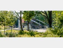 Moderner Bahnhof mit Glas- und Metallkonstruktionen, umgeben von Bäumen und dichter Vegetation. Zwei breite Treppenaufgänge führen zu einer erhöhten Plattform, während Fahrradständer und Wege im Vordergrund sichtbar sind.