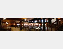 Großer Saal im Stil eines Western-Saloons mit Holzmöbeln, runden Tischen und Stühlen, hölzernen Balustraden und einer Empore. Deckenleuchten hängen von der Holzdecke, und rechts befindet sich eine Treppe mit geschnitztem Geländer.