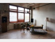 Industrie-ähnlicher Raum mit großer Fensterfront und Holzjalousien. Der Raum enthält eine Sitzgruppe mit einem grünen Polsterstuhl, einem antiken Sofa und einem niedrigen Tisch aus Holzpaletten, dekoriert mit Blumen; außerdem stehen Lautsprecher und eine Stehlampe im Raum.