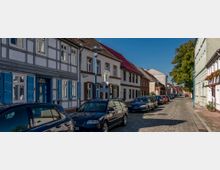 Gepflasterte Straße in einer Altstadt mit parkenden Autos links und rechts, flankiert von Fachwerk- und Putzhäusern mit bunten Fenstershuttern und roten Ziegeldächern; im Hintergrund ein Baum und blauer Himmel.