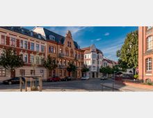 Straßenszene mit historischen Wohngebäuden im Stil der Gründerzeit, gekennzeichnet durch Backsteinfassaden und Mansarddächer. Im Vordergrund stehen Autos am Straßenrand und junge Bäume, rechts ist ein rotes Backsteingebäude mit großen Fenstern sichtbar.