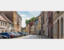 Eine gepflasterte Straße in einer Altstadt mit bunten historischen Gebäuden und Laternen. Links sind geparkte Autos, rechts befinden sich Arkaden mit Rundbögen.