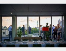 Eine Terrasse auf einem Dach mit Blick auf eine Stadt im Hintergrund, sichtbar durch große Glasfenster. Im Vordergrund ein gedeckter Tisch mit Gläsern und Besteck, im Außenbereich stehen Personen in kleinen Gruppen unter Lichterketten zusammen.
