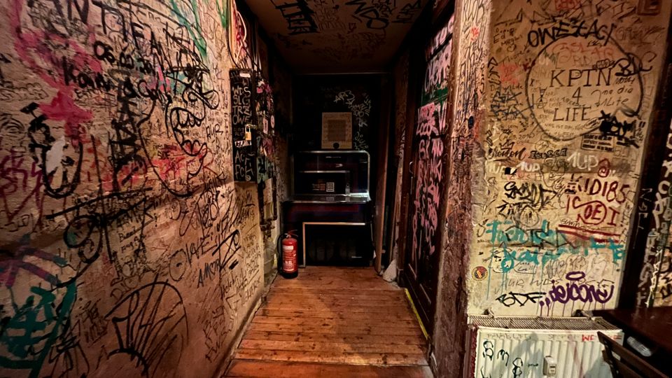 Schmaler Innenflur mit Holzfußboden, dessen Wände und Decke dicht mit Graffiti, Kritzeleien und Aufklebern bedeckt sind. Am Ende des Gangs stehen ein kleiner Tresen oder Automat und ein Feuerlöscher; rechts ist ein Heizkörper mit weiteren Markierungen zu sehen.