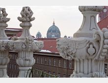 Blick auf die Kuppel des Berliner Doms zwischen zwei verzierten steinernen Balustradenelementen im Vordergrund, mit roten Ziegeldächern von umliegenden Gebäuden im Hintergrund.