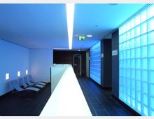 Moderner Innenraum eines Gebäudes mit bläulich beleuchteten Glasbausteinwänden, schwarzen Fliesen, einer Reihe von Liegestühlen auf der linken Seite und einer Sicherheitsbeleuchtung über einem Flur. Die Decke hat ein langes, eingelassenes Lichtband.