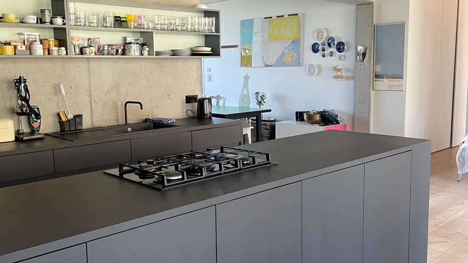 Moderne Küche mit einer großen, mattschwarzen Kücheninsel mit integriertem Gaskochfeld und einer schlichten Arbeitsplatte. Die Rückwand aus Sichtbeton enthält offene Regale mit Geschirr, und der Boden besteht aus hellem Holz.
