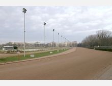 Rennbahn mit sandigem Untergrund, flankiert von hohen Straßenlampenreihen und weißen Zäunen. Im Hintergrund befinden sich mehrstöckige Gebäude und eine Baumreihe unter bewölktem Himmel.