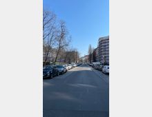 Straßenansicht einer Wohngegend mit parkenden Autos auf beiden Seiten. Rechts ist ein Gerüst an einem Gebäude zu sehen, während links mehrere Bäume mit kahlen Ästen stehen; blauer Himmel im Hintergrund.