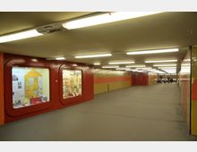 Unterirdischer Fußgängertunnel mit gelben und roten Wandfliesen, beleuchtet durch zahlreiche Leuchtstofflampen an der Decke. Auf der linken Seite befinden sich zwei Schaufenster mit Ausstellungen, rechts eine glatte Wand, und im Hintergrund sind mehrere Personen erkennbar.