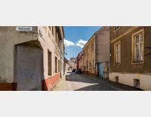 Schmale Altstadtstraße mit Kopfsteinpflaster, umgeben von verwitterten Gebäuden mit teilweise abblätterndem Putz; ein Straßenschild mit der Aufschrift „Wallstraße“ ist sichtbar. Autos parken entlang der rechten Seite, während im Hintergrund weitere Gebäude und ein blauer Himmel mit Wolken zu sehen sind.