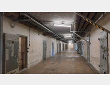 Langer, schmaler Korridor in einem alten Gefängnis oder Bunker mit massiven Metalltüren auf beiden Seiten. Die Wände sind abgenutzt, und an der Decke verlaufen freiliegende Rohre und Leitungen, beleuchtet von Neonröhren.