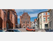 Straßenszene in einer Altstadt mit gepflastertem Platz und historischen Backsteingebäuden, darunter ein gotisches Rathaus im Zentrum mit spitzem Giebel. Links ein Kirchengebäude mit großen Spitzbogenfenstern, rechts helle Fassaden modernerer Wohnhäuser.