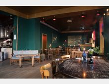 Gemütliches Café mit dunkelgrünen Wänden, Holzmöbeln und Kerzenlicht auf den Tischen. Im Vordergrund stehen rustikale Holztische mit Blumenvasen, im Hintergrund befindet sich eine kleine Sitzecke und eine Theke.