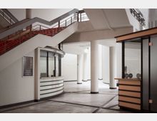 Modernes Foyer mit weißer Inneneinrichtung, roten Treppengeländern und einer Ticketkasse mit abgerundeter Glasfront. Sichtbare Designelemente sind schlanke Säulen, ein heller Boden und minimalistische Formen.