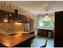 Küche im Retro-Stil mit gemusterten Fliesen an den Wänden, braunen Holzschränken und einer Theke. Ein großes Fenster mit weißen Vorhängen bietet Blick ins Grüne, und eine Hängelampe beleuchtet den Raum.