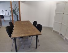 Ein moderner Besprechungsraum mit einem rechteckigen Holztisch, umgeben von schwarzen Stühlen. Im Hintergrund ein Glastrennwand mit Blick auf ein angrenzendes Büro mit Schreibtisch, Lampe und Topfpflanze; rechts ein weißer Raumteiler.