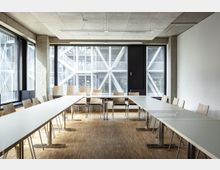 Ein moderner Besprechungsraum mit langen, weißen Tischen in U-Form, umgeben von Holzstühlen. Der Raum hat große Fenster mit Blick auf eine Außenfassade aus Metallstreben und eine Holzbodenfläche.