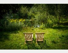 Zwei Holzstühle stehen auf einer grünen Wiese, die von hohen Gräsern, Büschen und Bäumen umgeben ist, im Hintergrund ist ein kleiner Teich mit Wasserpflanzen sichtbar.
