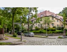 Kreuzung einer gepflasterten Wohnstraße mit anliegenden Wegweisern. Im Hintergrund stehen beige Mehrfamilienhäuser mit roten Ziegeldächern und großen Fenstern, umgeben von Bäumen und gepflegten Grünanlagen.