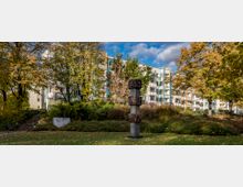 Wohnanlage mit moderner Architektur, umgeben von Bäumen und einem grünen Park. Im Vordergrund steht eine dekorative Skulptur mit abstrakten Mustern auf einem säulenartigen Sockel.