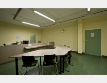Ein schlicht eingerichteter Büroraum mit gelben Wänden und grünem Bodenbelag. Zu sehen sind ein Empfangstresen mit Monitor und Stühlen sowie ein großer, dreiecksförmiger Tisch mit mehreren Stühlen; an der Wand eine Uhr und eine grüne Tür.