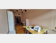 Schmales Büro mit Holzfußboden, hellen Wänden und moderner Deckenleuchte; links stehen hohe weiße Schränke, rechts ein großer Schreibtisch mit Ablagen. Im Hintergrund sind ein weiterer Arbeitsplatz, ein Regal und eine offene Tür zu einem Flur zu sehen.