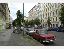 Straßenszene in einer europäischen Stadt mit Altbauten, die mehrere Stockwerke hoch und in bunten Farben gestrichen sind. Am Straßenrand sind Bäume, parkende Autos und ein Gehweg mit Kopfsteinpflaster zu sehen.