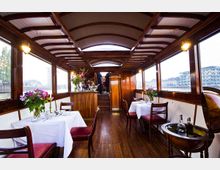 Innenraum eines eleganten Bootes mit Holzausstattung, einem gewölbten, mit Querstreben versehenen Dach und großen Fenstern, die den Blick auf die umliegenden Gebäude ermöglichen. Der Raum ist mit weiß gedeckten Tischen, roten Polsterstühlen und Blumenarrangements dekoriert, im Hintergrund befindet sich eine kleine Bar.