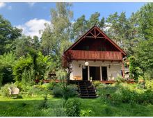 Ein zweistöckiges Landhaus mit rotem Holzgiebel und weißer Fassade, umgeben von dichtem Wald. Vor dem Haus befindet sich ein Garten mit verschiedenen Pflanzen, einer Rasenfläche und Gartenmöbeln.