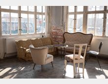 Ein heller Raum mit großen, rustikalen Fenstern und auf alt getrimmtem Interieur, bestehend aus gepolsterten Sofas, einem kleinen runden Tisch und verschiedenen Stühlen, auf einem Betonboden. Die Backsteinwand im Hintergrund und sichtbare Heizkörper unter den Fenstern betonen den industriellen Charakter.
