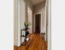 Schmaler Flur mit Holzdielenboden, hellen Wänden und weißen Türrahmen. Links steht ein schwarzes offenes Regal mit Deko-Elementen, rechts befindet sich eine gepolsterte Sitzbank.