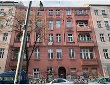 Mehrstöckiges Altbauwohnhaus in einer städtischen Umgebung, mit rosafarbener Fassade und verzierten Fenstern sowie Balkonen. Im unteren Bereich sind die Wände mit Graffiti bedeckt, und vor dem Gebäude parken Autos; ein kahler Baum und eine Straßenlaterne ragen in den Vordergrund.