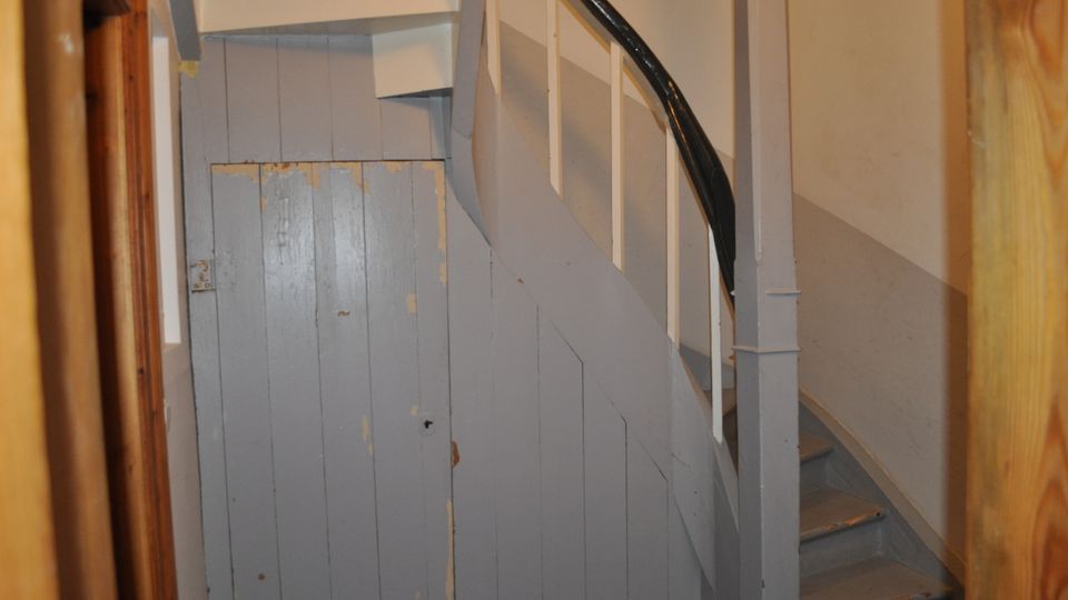 Eine enge Innentreppe aus Holz mit abgenutztem grauen Anstrich und schwarzem, glänzend lackiertem Handlauf. Rechts von der Treppe befindet sich eine Wand, links ein graues Holztür-Panel mit sichtbaren Gebrauchsspuren.