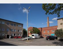 Mehrstöckige Wohngebäude aus Putz- und Backstein in einem Innenhof, mit geparkten Autos und einem Lieferwagen. In der Mitte ein hoher Laternenmast, im Hintergrund ein Backsteingebäude mit einem Schornstein.