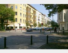 Straßenkreuzung in einem städtischen Wohngebiet mit mehrstöckigen, hell gestrichenen Altbauten. Gehwege mit Bäumen, parkende Autos, Verkehrsschilder und ein Tempo-30-Schild sind sichtbar; im Hintergrund Fußgänger und ein Fahrradständer.