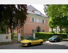 Ein rosa Gebäude mit Satteldach und weißen Fensterläden, umgeben von einer Hecke und einem Metallzaun. Vor dem Haus parken ein gelbes Cabrio und ein grünes Auto; Bäume säumen die Straße im Vordergrund.