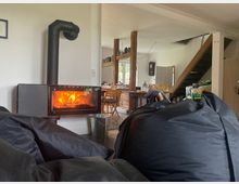 Gemütliches Wohnzimmer mit einem modernen, schwarzen Kaminofen, der befeuert ist. Im Hintergrund Holzstühle, ein Esstisch mit Geschirr und eine offene Holztreppe, die ins obere Stockwerk führt.