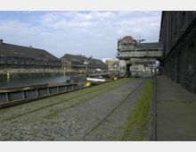 Historischer Hafenbereich mit alten Lagerhäusern aus Backstein entlang eines Kanals. Im Vordergrund sind Kopfsteinpflaster, Gleise und Anlegestellen für Schiffe sowie teils modernisierte Hafenstrukturen erkennbar.