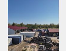 Gewerbefläche mit mehreren Containern in verschiedenen Farben, offenen Lagerhallen mit roten Blechdächern und gestapeltem Holz im Vordergrund; im Hintergrund Bäume und blauer Himmel.