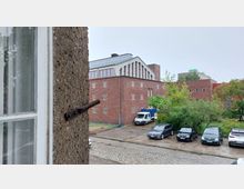 Blick aus einem Fenster auf ein modernes Backsteingebäude mit flachem Dach und großen Fenstern an der oberen Fassade, umgeben von Bäumen und Sträuchern. Im Vordergrund sind eine gepflasterte Zufahrt und mehrere geparkte Fahrzeuge zu sehen.