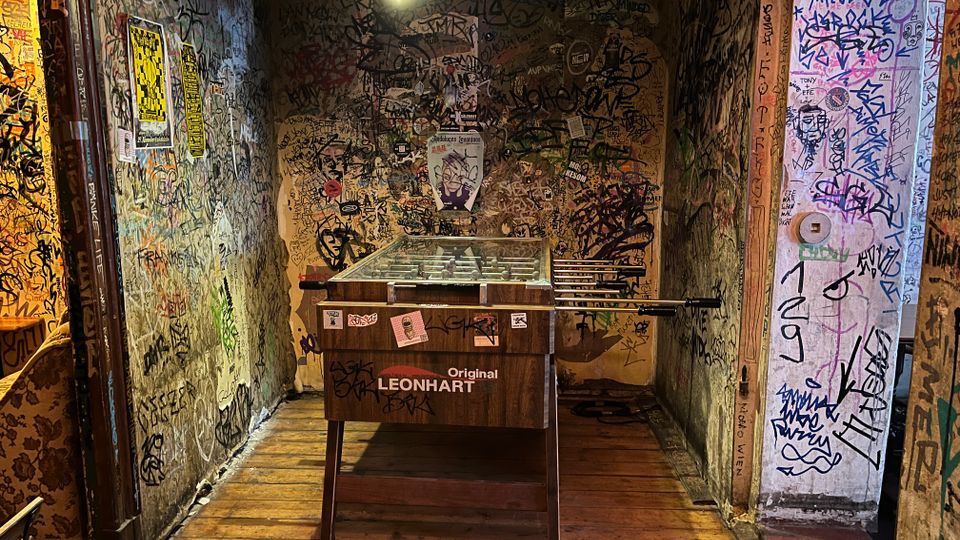 Schmaler Innenraum wie ein Flur in einer Bar oder einem Klub, mit Holzboden und einer Deckenlampe über einem zentral stehenden Kickertisch. Die Wände und Türrahmen sind dicht mit Graffiti, Stickern und Postern bedeckt.