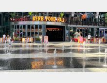 Eine moderne Food-Hall mit der beleuchteten Schrift "KERB FOOD HALL" und einer Glasfassade, umgeben von roten Tischen und Stühlen im Außenbereich. Im Vordergrund befindet sich ein Brunnen mit sprudelnden Wasserfontänen auf einer gepflasterten Fläche.