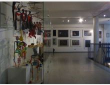Innenraum einer Kunstgalerie mit hell beleuchtetem Ausstellungsbereich. Links im Bild befinden sich bunte Skulpturen und Puppen hinter einer Glasvitrine, rechts an der Wand sind gerahmte Gemälde in mehreren Reihen aufgehängt.