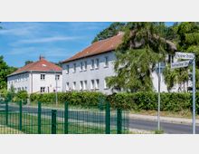 Straßenszene mit zwei weißen, zweistöckigen Wohngebäuden mit roten Ziegeldächern, umgeben von grünen Hecken und Bäumen. Im Vordergrund führt ein Gehweg entlang einer Straße, und Straßenschilder markieren die Kreuzung von "Ruppiner Straße" und "Fehrbelliner Straße".