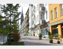 Straßenszene mit mehrstöckigen Wohn- und Geschäftshäusern, teils in weißer und gelber Fassade, mit Balkonen und Schaufenstern im Erdgeschoss; im Hintergrund ragt ein Kirchturm in die Höhe, und links säumen Bäume die Straße. Ein gelbes Fahrzeug parkt auf der linken Seite entlang des Bürgersteigs.