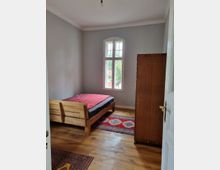 Ein einfach eingerichtetes Schlafzimmer mit Holzboden, einem Holzbett mit roter Bettdecke, einem hohen Kleiderschrank aus Holz und einer großen Fensterfront mit weißem Rahmen, die Tageslicht hereinlässt. Zwei farbige Teppiche liegen auf dem Boden.