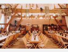 Großer, rustikaler Veranstaltungsraum mit Holzbalkenkonstruktion, langen Holztischen und Klappstühlen, dekoriert mit Blumen, getrockneten Pflanzen und hängenden Papierlampen. Im Hintergrund befinden sich Holzmöbel, warme Beleuchtung und Dekorationen im Vintage-Stil.